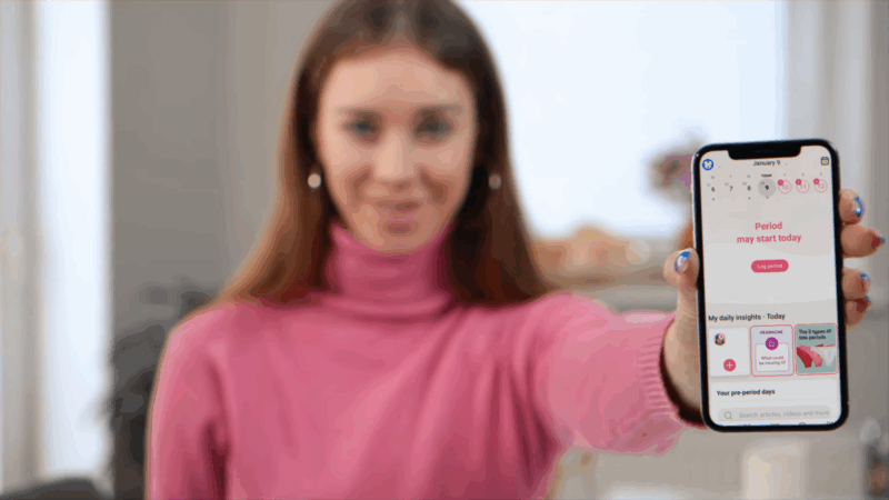 A woman holds up her smartphone showing a period tracking app with daily insights and cycle details