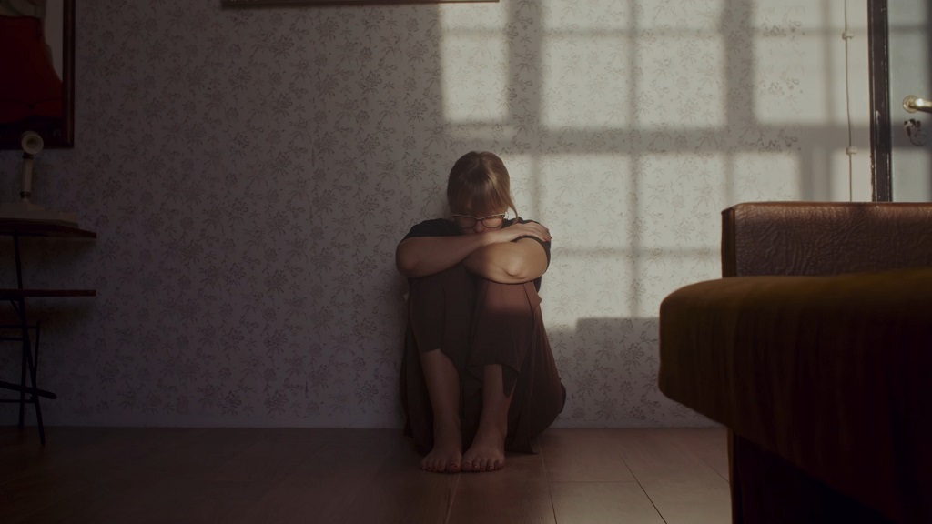 Woman sitting alone on the floor in a dimly lit room, hugging her knees, conveying feelings of stress or sadness