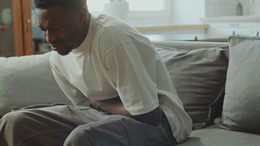 Man sitting on a couch, clutching his stomach in visible discomfort, suggesting digestive pain or gut issues